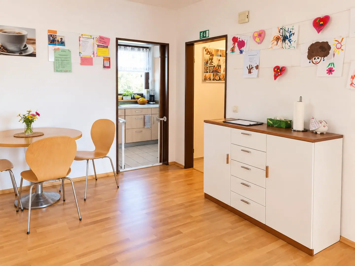 Café-Ecke mit Tisch und Blumen, Blick in die kleine Küche; an der Wand hängen Kinderbilder und gebastelte Herzen.