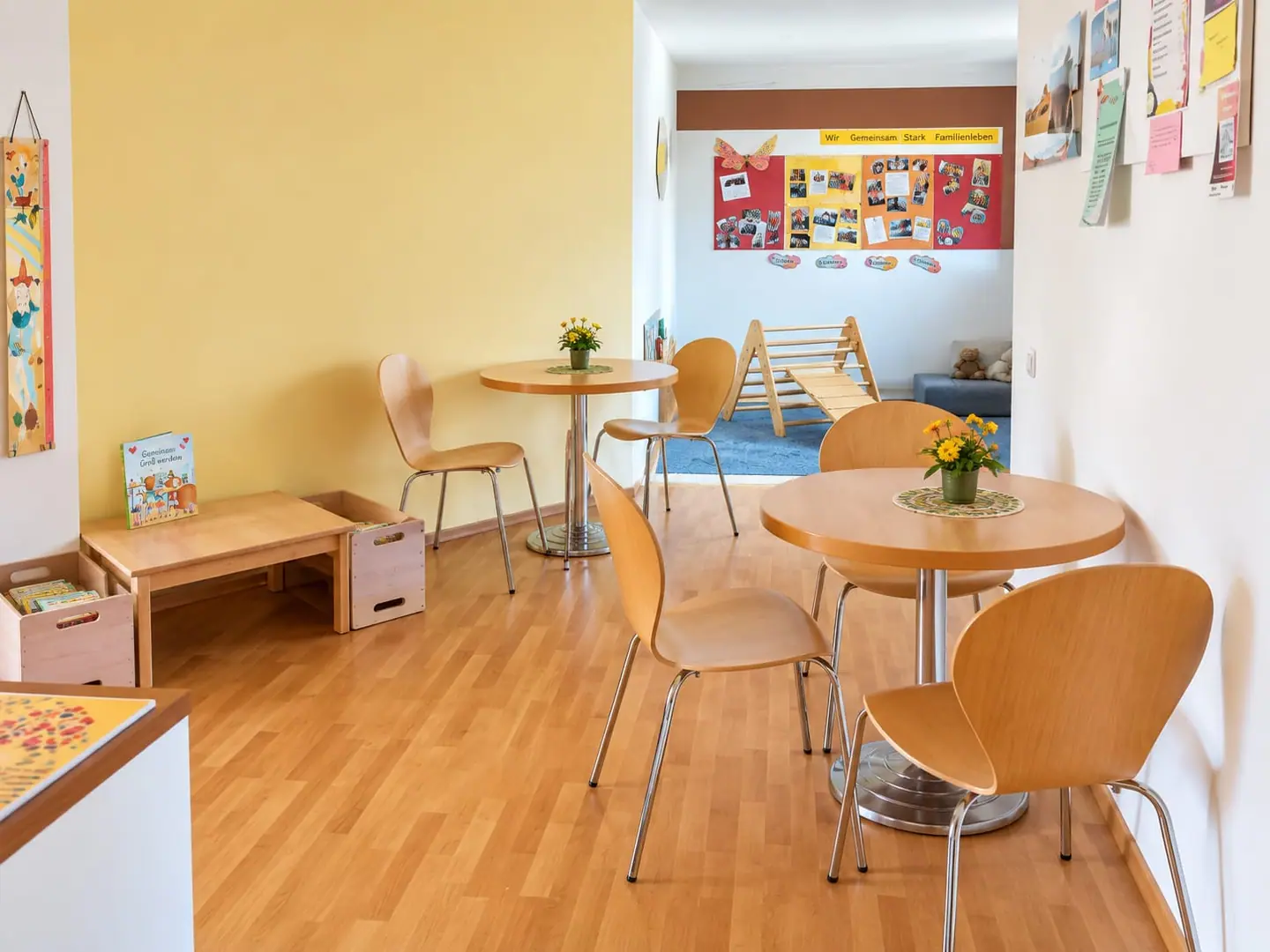 Sitzecke mit gelber Wand, Holztischen und Blumen; im Hintergrund eine Spielfläche mit Kletterdreieck und Plakat „Wir gemeinsam stark – Familienleben“.