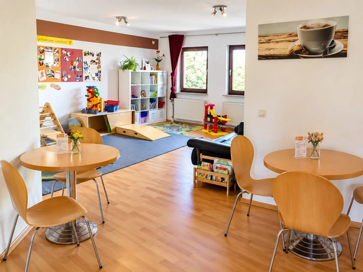 Familiencafé mit runden Holztischen und Blumen, dahinter eine Spielecke mit Kletterdreieck, Rutsche, Bücherregal und Spielteppich.